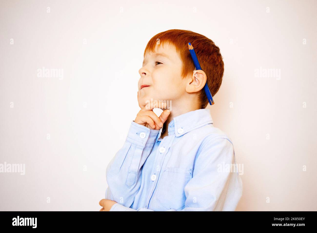 a little red-haired boy cheerful and thoughtful in a shirt with a pencil on a white background Stock Photo