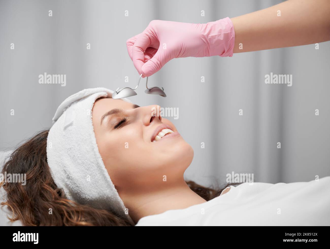 Close up of woman's face who lying with closed eyes at reception at beautician. Cropped view of hand in pink rubber glove keeping metal eye shield from laservision on short distance from face. Stock Photo