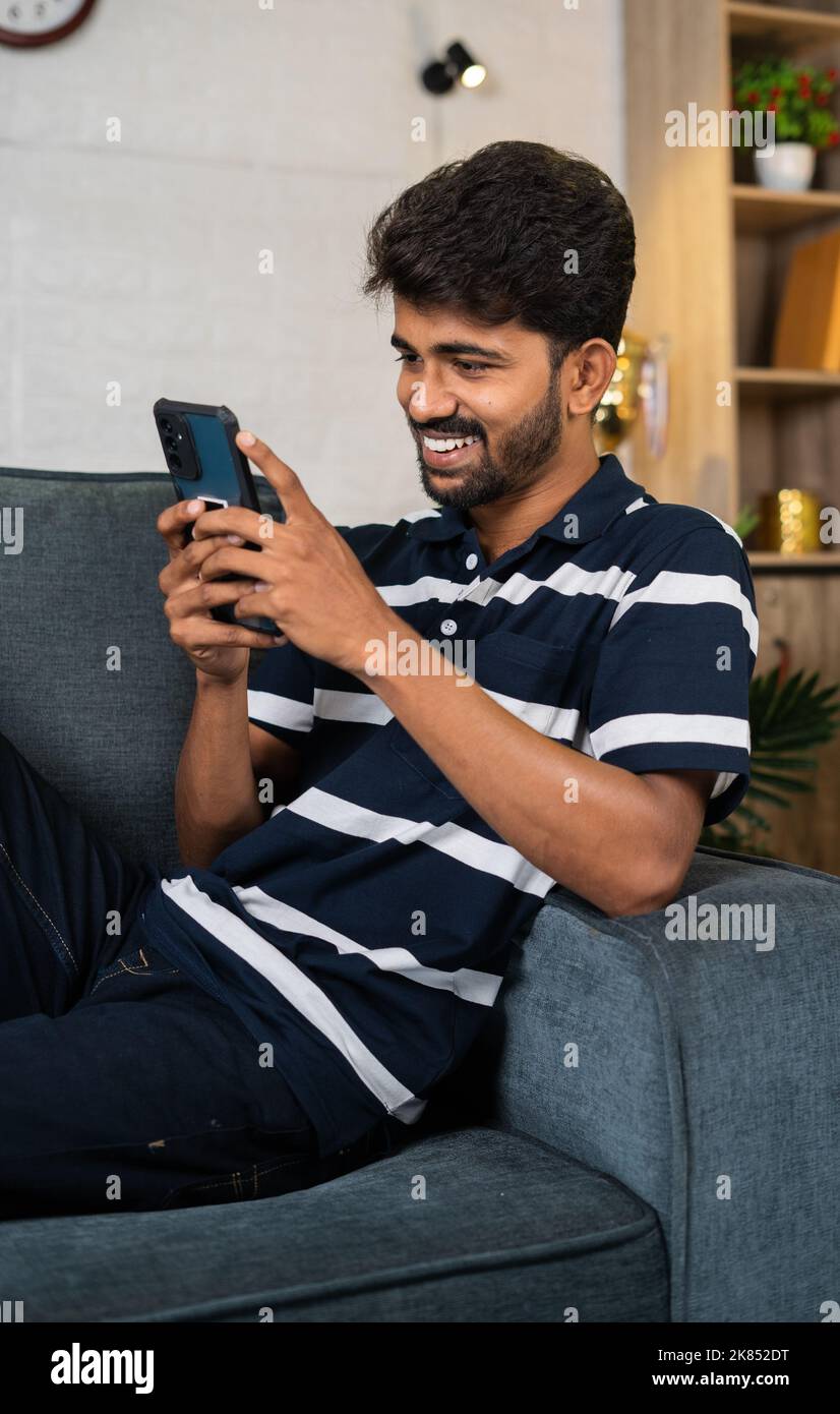 vertical shot of Happy smiling young man using mobile phone while sitting on sofa at home - concept of social media sharing, internet and relaxation Stock Photo