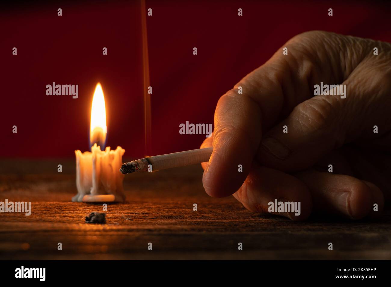 old woman holding a cigarette by candlelight in the dark at home at the table, smoking at home in the dark, nicotine and bad habit, cigarettes and can Stock Photo