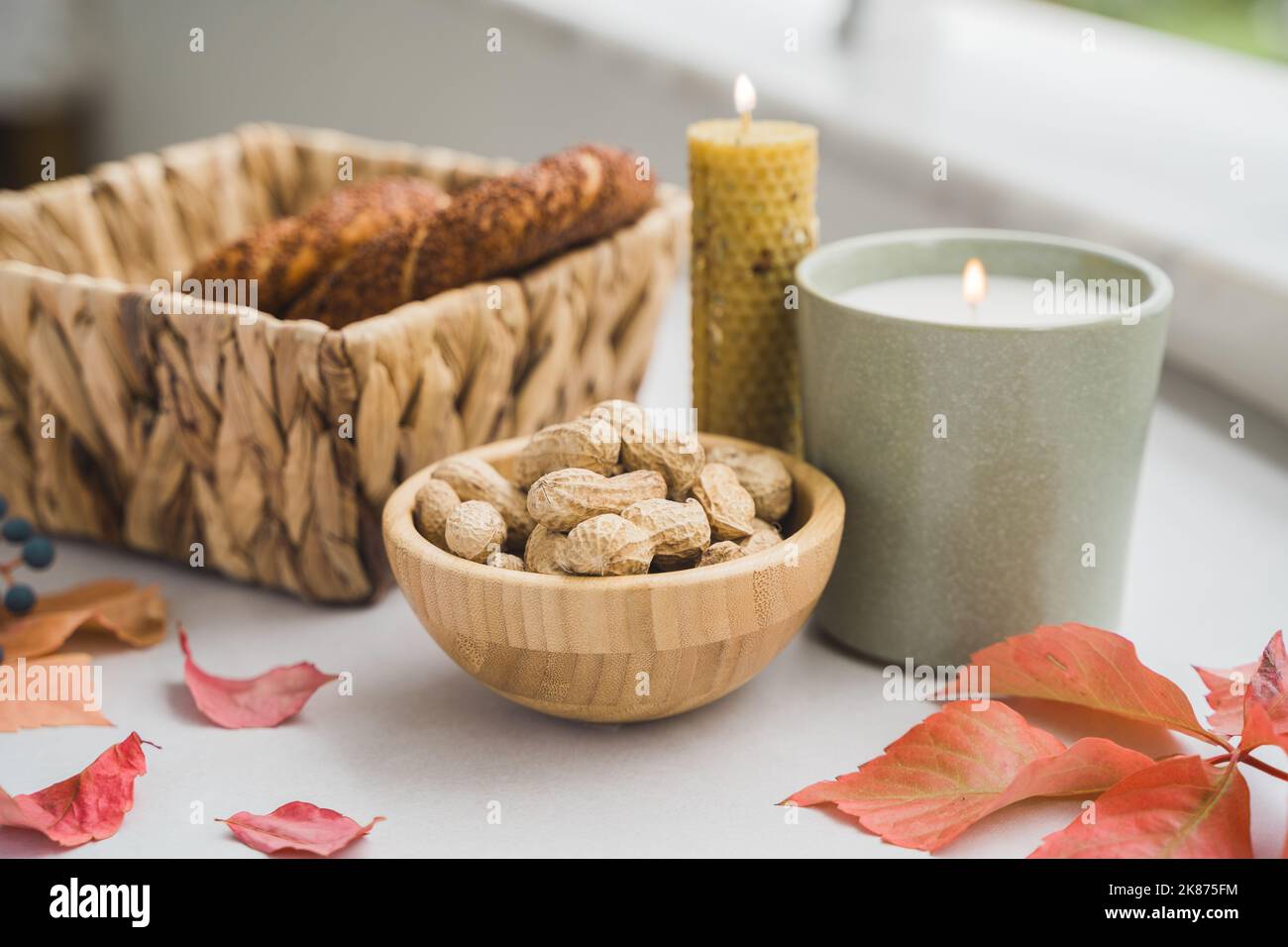 Still life candle flame, peanut, fallen leaves, wicker basket, bagel on a table, home decor in a cozy house. Autumn weekend concept. Stock Photo