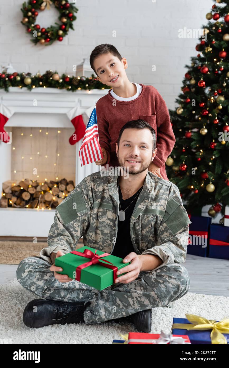 happy boy with usa flag and military man with christmas present looking at camera at home Stock Photo
