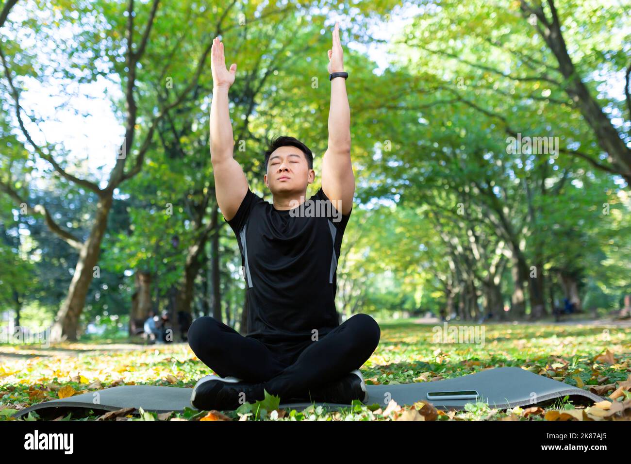 Asian man in the park on a sports mat sits in the lotus position, sportsman performs breathing exercises on a sunny day, man in a sports suit rests after physical and fitness exercises Stock Photo