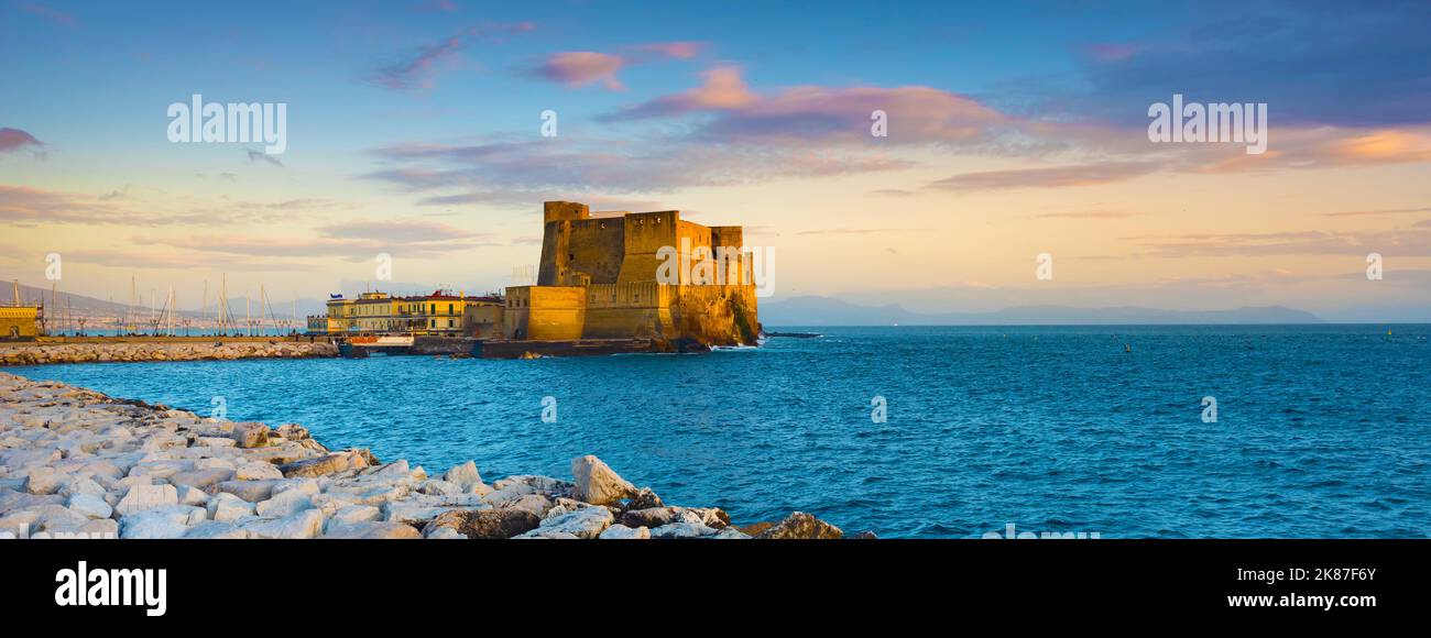 Naples, Italy. Castel dell'Ovo with breakwater rocks in the foreground and with a beautiful sunset sky. Banner Header. Stock Photo