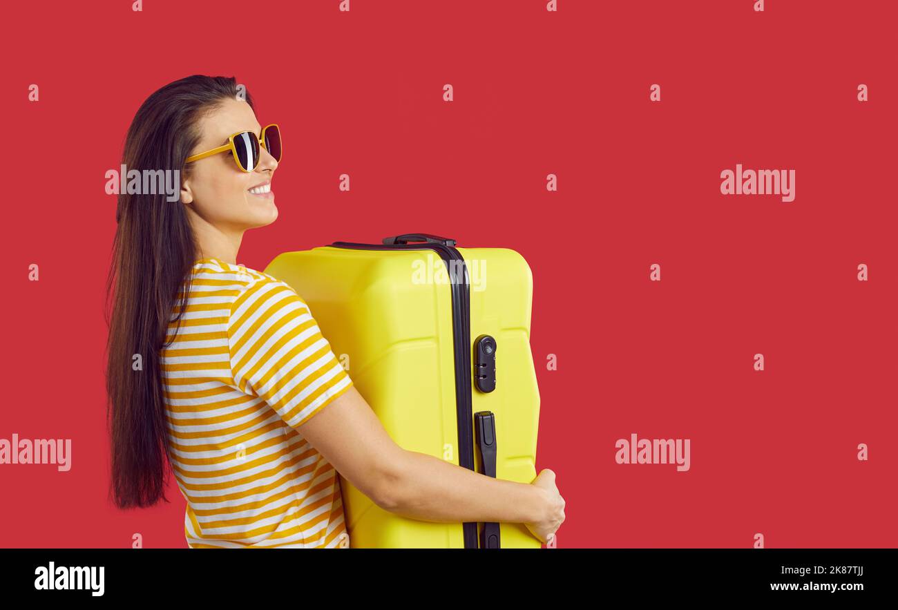 Joyful young woman with yellow suitcase on red background ready for summer vacation. Stock Photo