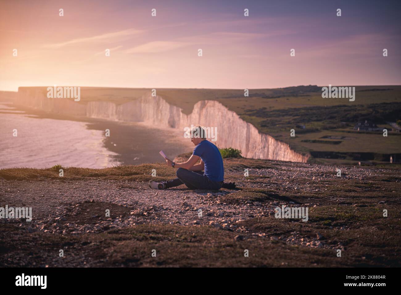 A man sitting on a rocky cliff watching the Seven Sisters cliffs in Eastbourne, United Kingdom at sunset Stock Photo