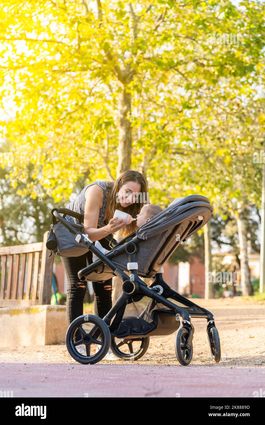 Mother feeding her son with a milk bottle in the park inside the baby stroller Stock Photo