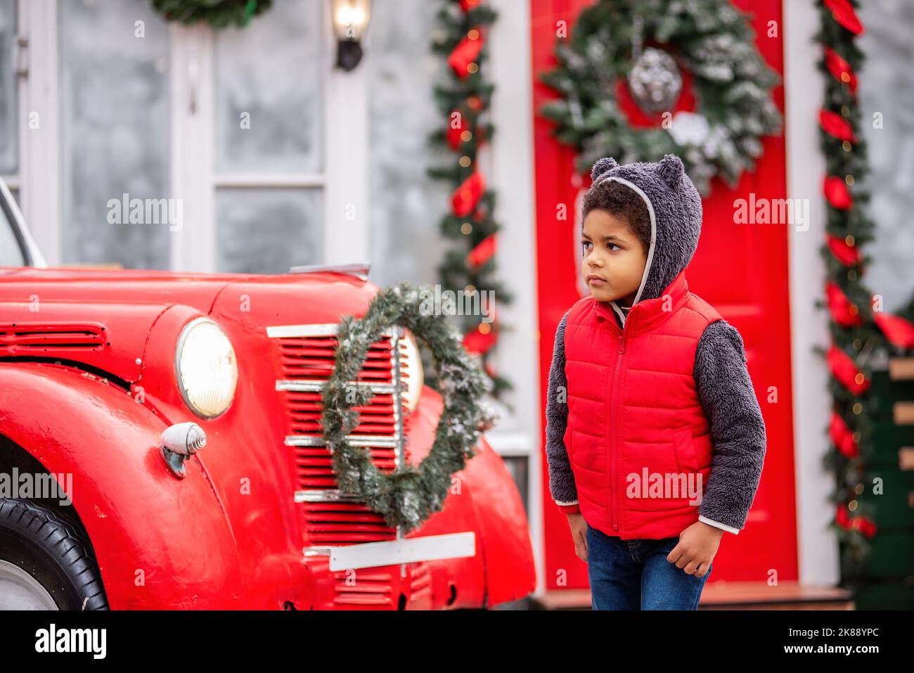 Close-up portrait Little African American boy in a bear costume, a red winter vest on the background of a wooden decorated door with a Christmas wreat Stock Photo