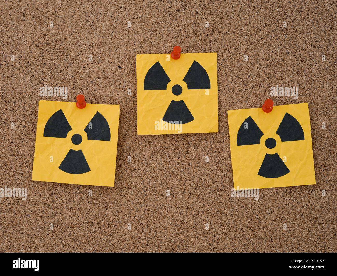 Three yellow paper notes with radiation warning signs on them pinned to a corkboard. Close up. Stock Photo