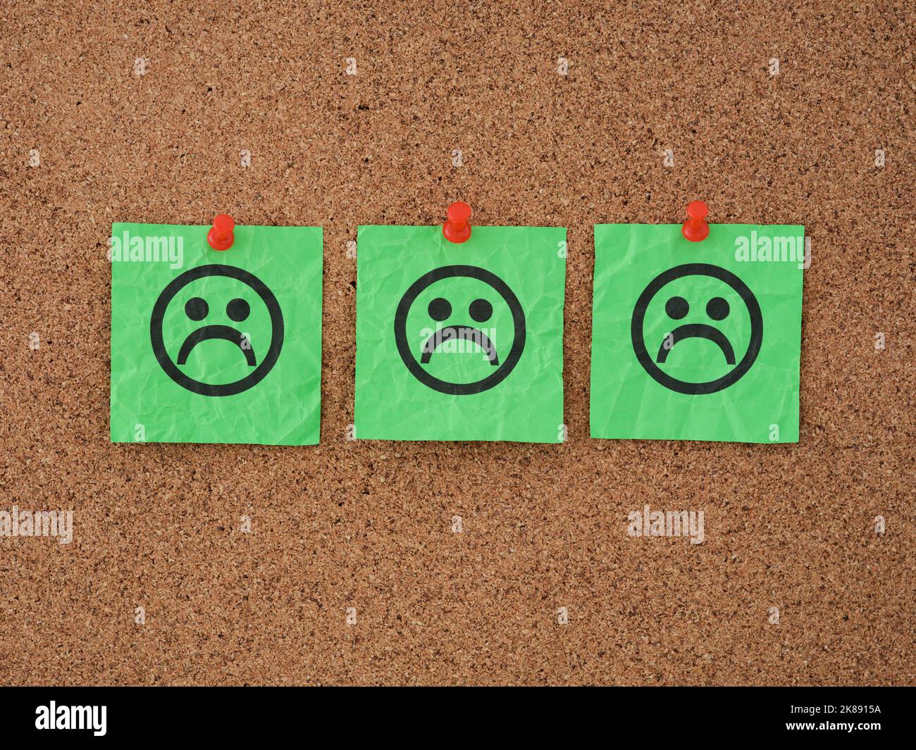 Three green paper notes with sad faces on them pinned to a corkboard. Close up. Stock Photo