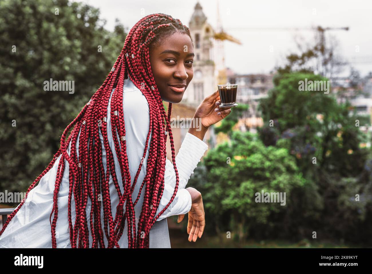 A portrait of a ravishing youthful black female with long red braids enjoying her morning espresso coffee while leaning against the fencing of a balco Stock Photo