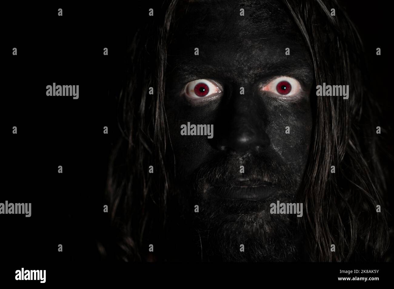 A scary face of a man in the dark. Long hair, black face and red eyes. Low light with dark background. Horror concept. Stock Photo