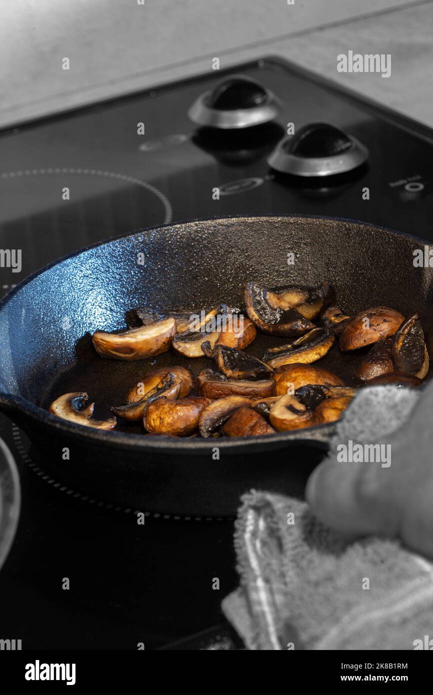 Frying chestnut mushrooms, with a man holding a cast iron frying pan with a tea towel, on an electric hob stove Stock Photo