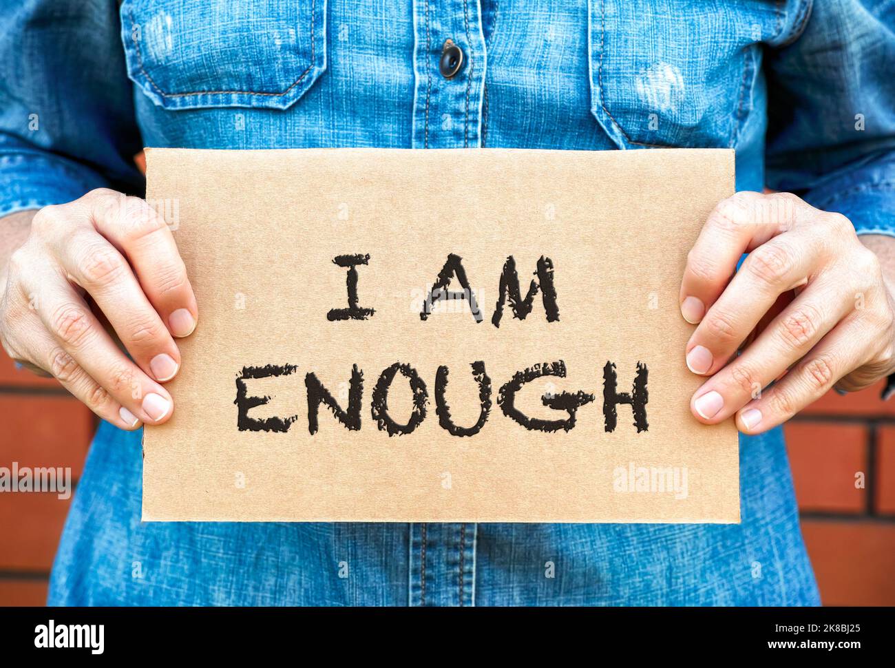 Woman hands holding a piece of a cardboard with words I am Enough against brick wall background. Stock Photo