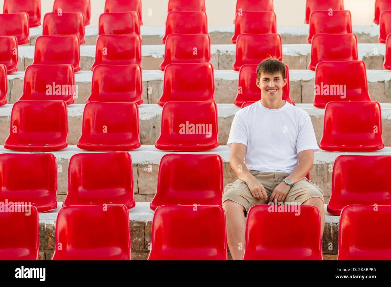 Wide smiling man with dental braces is sitting in deserted audience with bright red seats. Absence of people in open-air amphitheater. Stock Photo