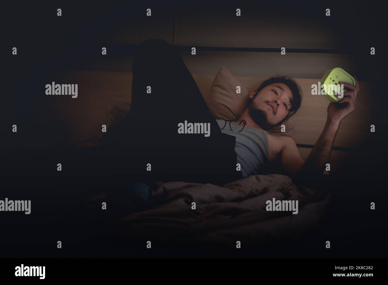Can't sleep at nighttime concept, Asian young Man holding and looking at Alarm Clock at night on the bed. Stock Photo