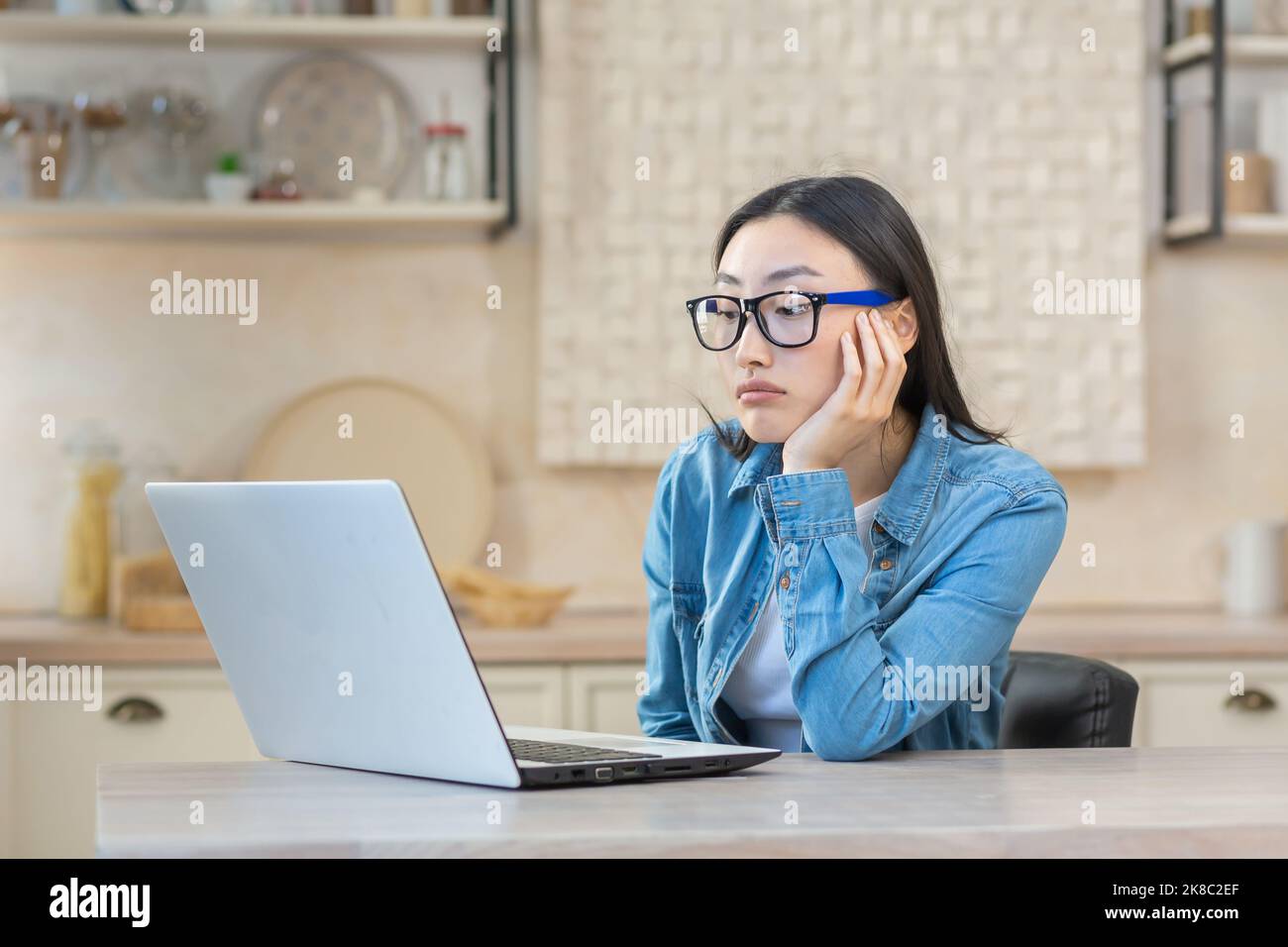 Tired young Asian woman freelancer sitting at a table with a laptop, bored, unable to start work. Works remotely at home during quarantine. Stock Photo