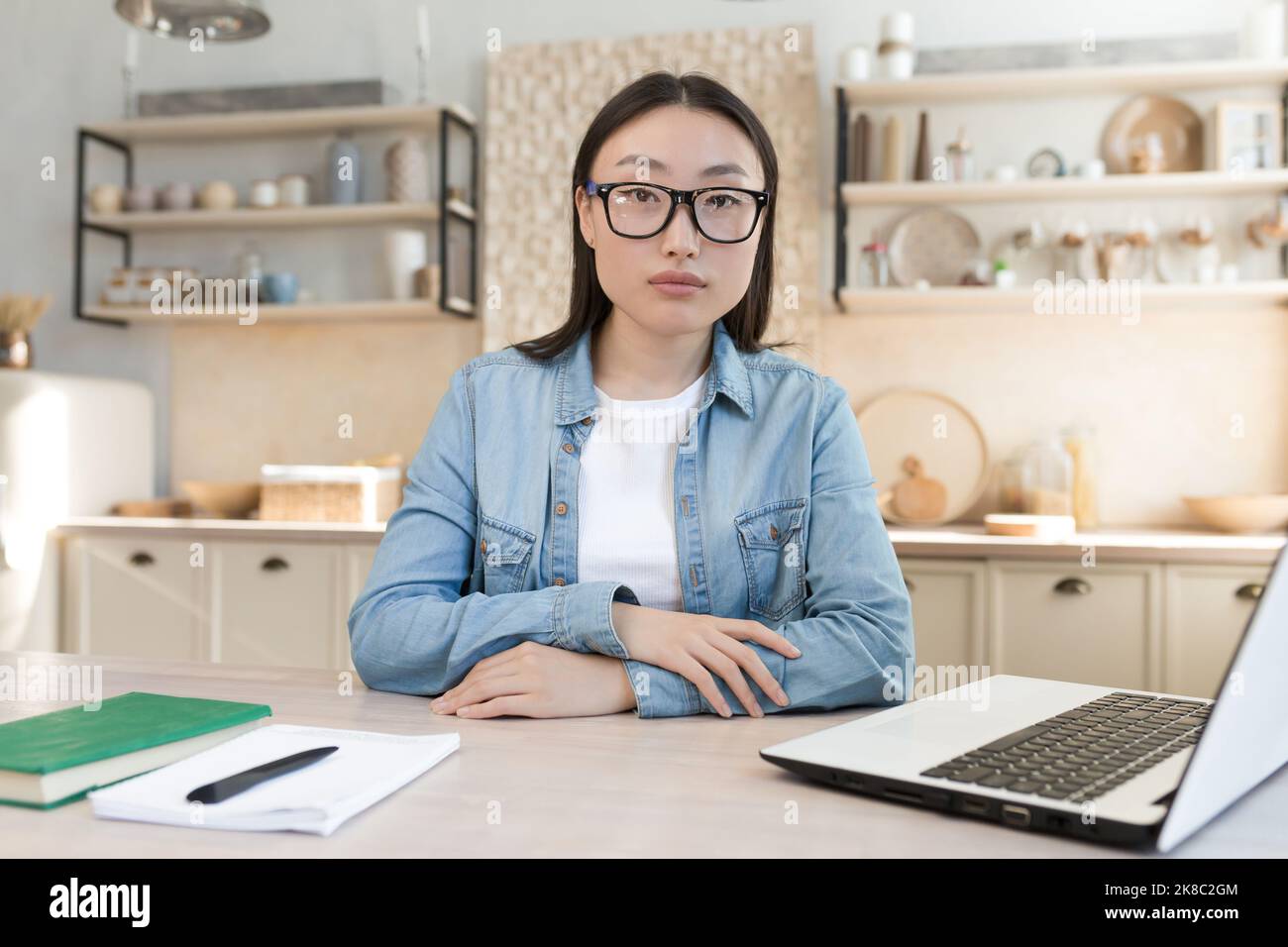 Portrait of a young Asian businesswoman working at home and conducting online interviews from a laptop. A serious, purposeful woman is sitting at the table, looking at the camera. Stock Photo