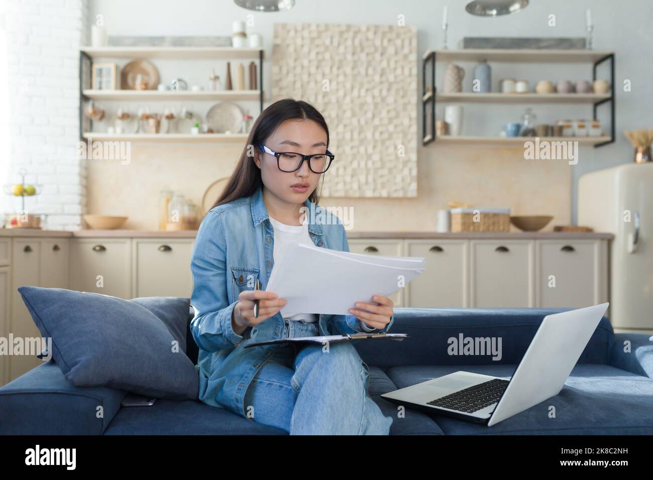 Young beautiful creative woman Asian writer, journalist. He sits at home on the sofa with a laptop and writes a book, an essay, a script, an article. Holds paper and pen. Stock Photo