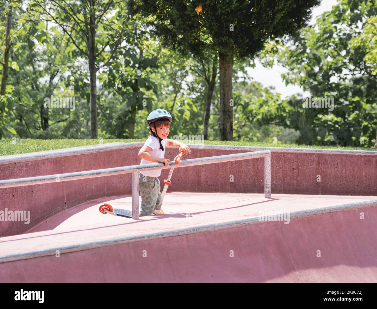 Little boy rides kick scooter in skate park. Special concrete bowl structures in urban park. Training to skate at summer. Stock Photo