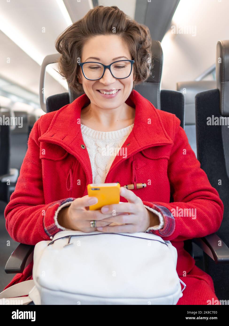 Smiling woman in red duffle coat texting on smartphone in suburban train. Travel by land vehicle. Stock Photo