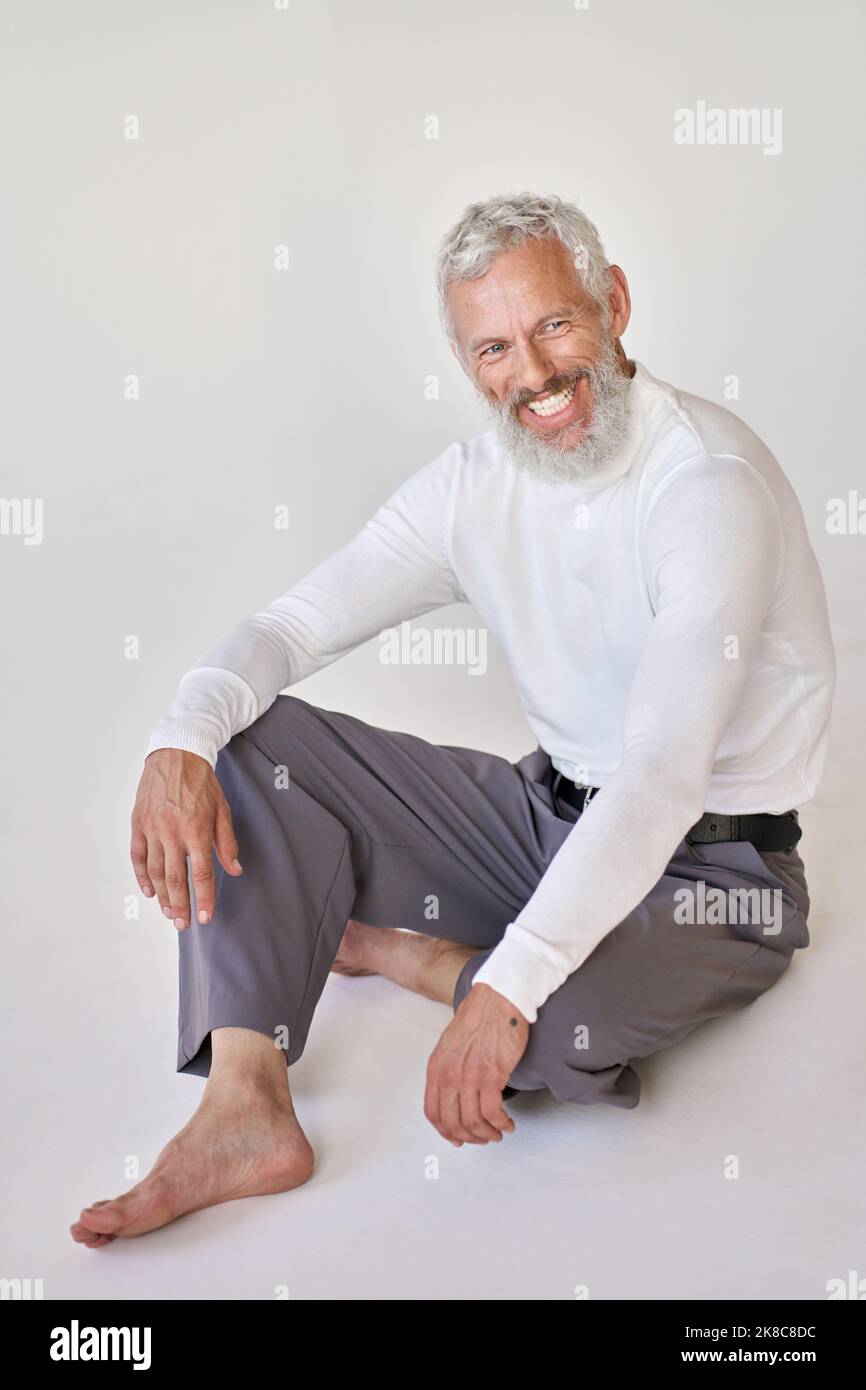 Smiling bearded senior older man sitting on floor at white wall. Stock Photo
