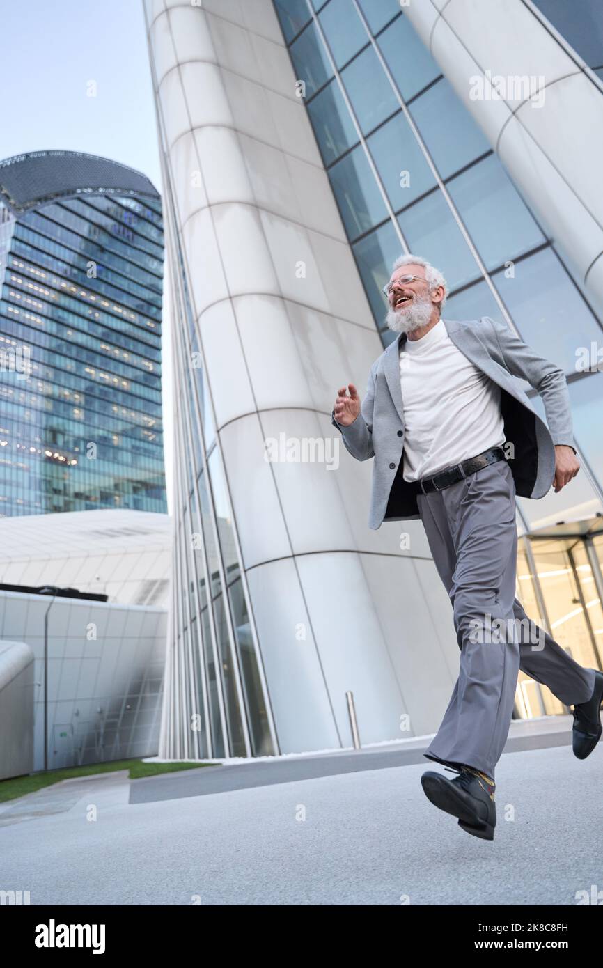 Happy stylish old business man wears suit running outdoor in big city street. Stock Photo
