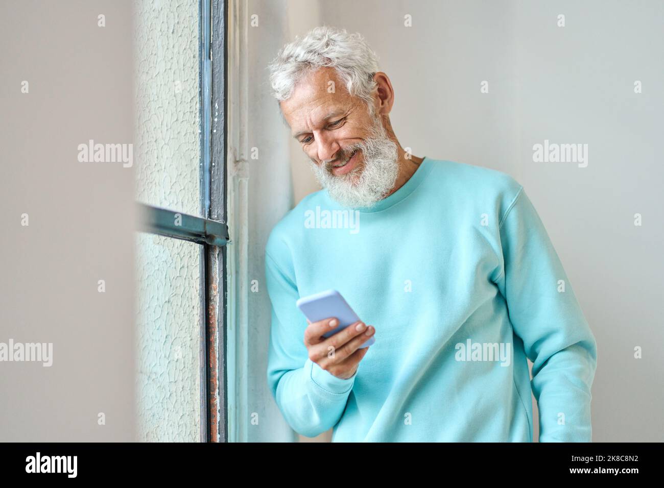 Smiling old bearded hipster man using mobile phone standing at window. Stock Photo