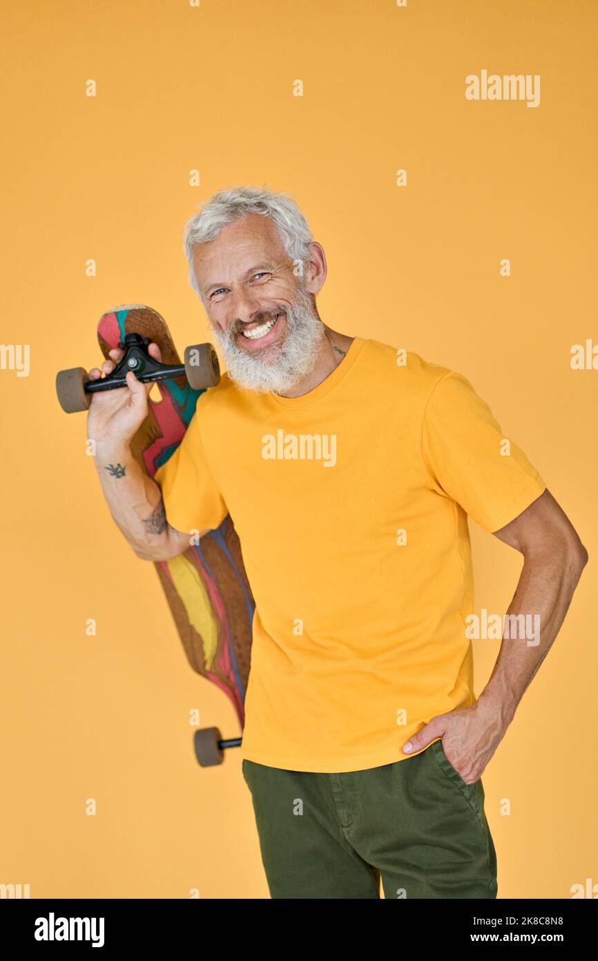 Happy bearded older man skater holding skateboard isolated on yellow. Stock Photo