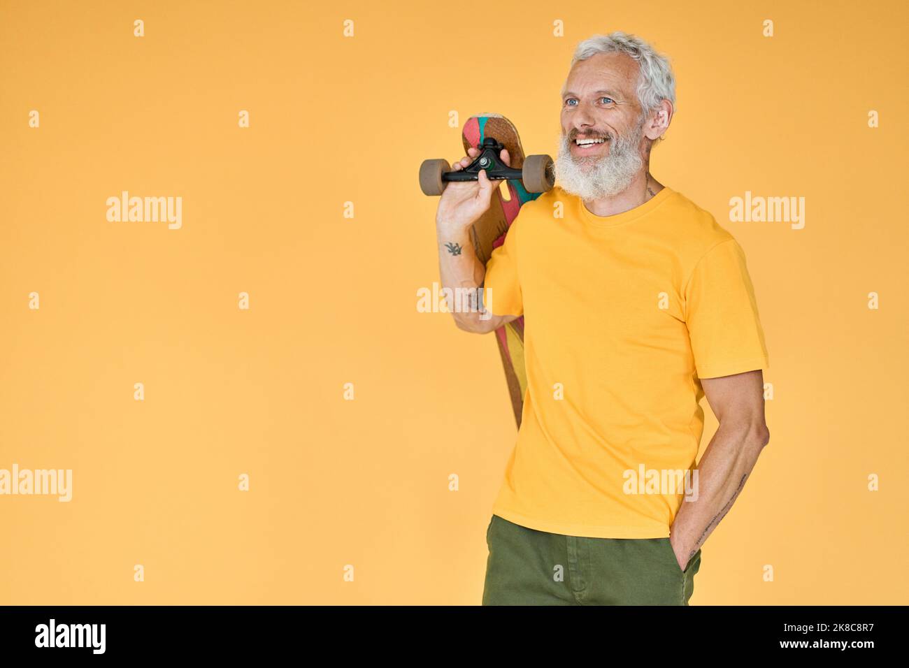 Happy bearded old man skater holding skateboard standing isolated on yellow. Stock Photo