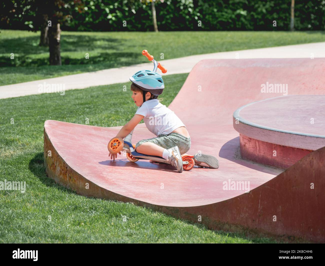 Little boy fell off kick scooter while riding in skate park. Special concrete bowl structures in urban park. Training to skate at summer. Stock Photo