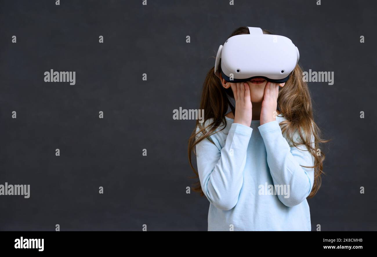 Kid in metaverse, child uses virtual reality headset for new experience. Surprised little girl hands up to face looking in VR glasses, young person pl Stock Photo