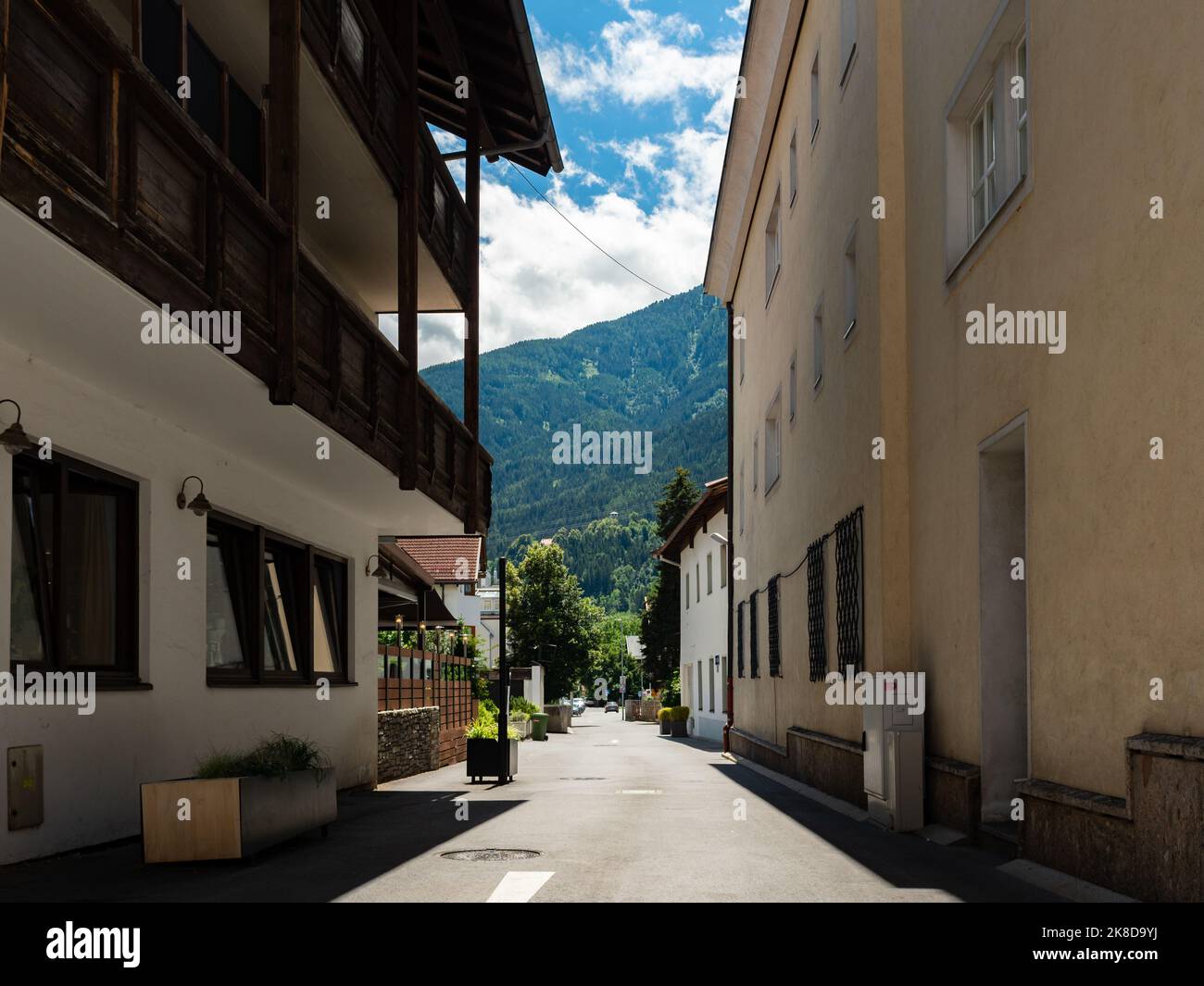 Narrow street in Tirol with buildings on the sides. Huge mountains are in the front. A forest with coniferous trees is part of the nature. Stock Photo