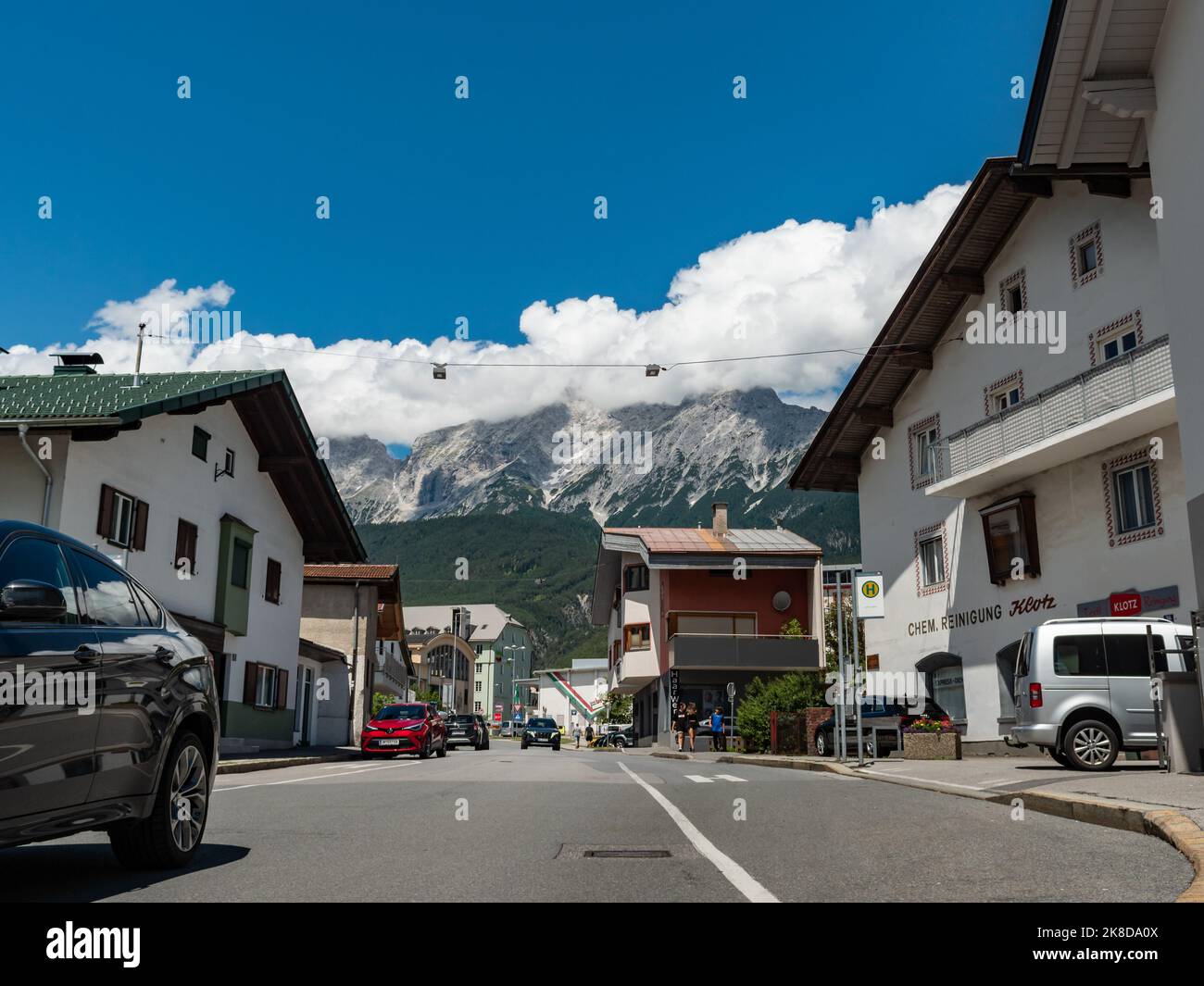 Street view in Tirol. People and cars are moving in the town. The road is going up steep and mountains of the Alps are in the background. White clouds Stock Photo