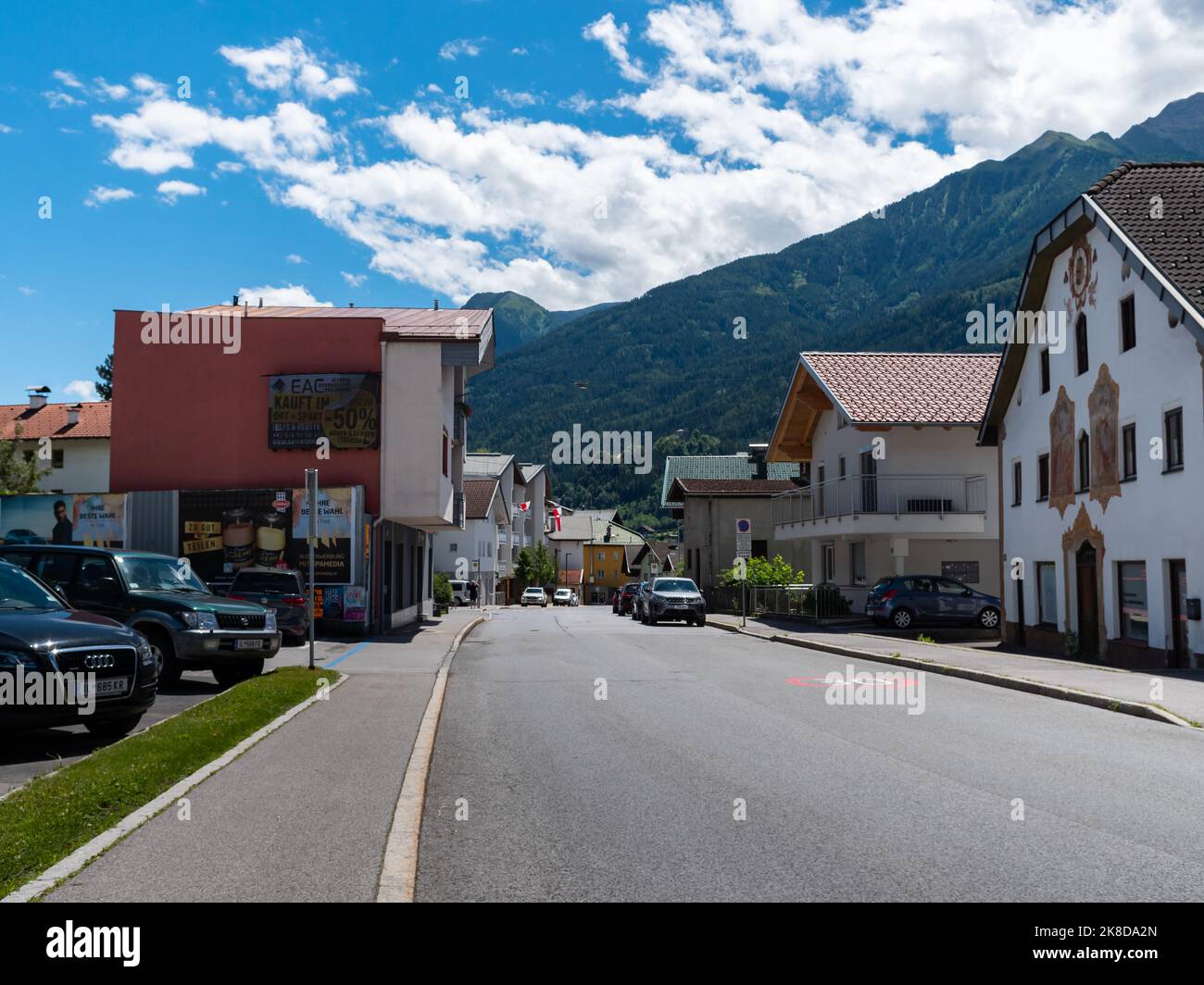 Street and residential buildings in Tirol. Cars are parking on the road. Mountains of the Alps are in the background and white clouds are on the sky. Stock Photo