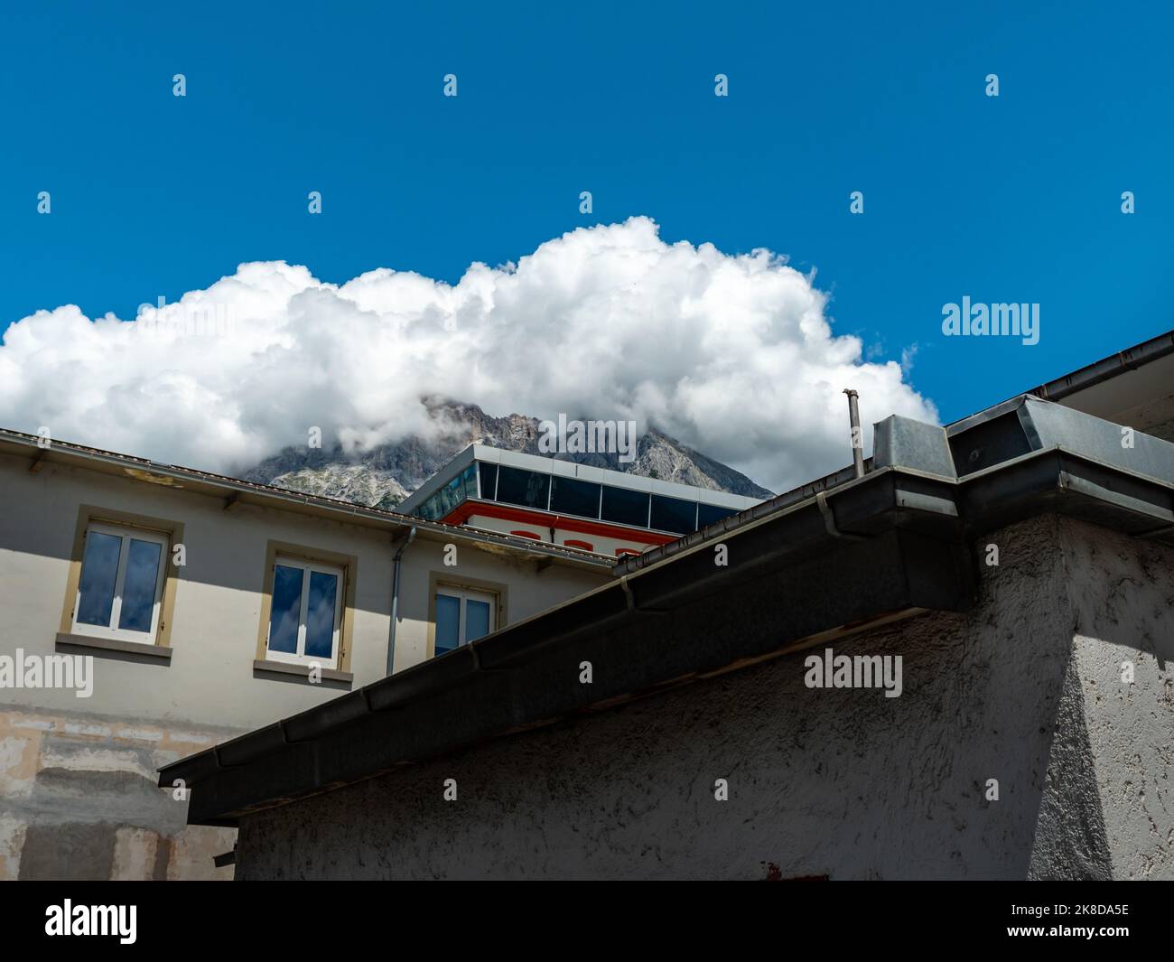 Buildings in front of the huge alpine mountain peak in Tirol. White clouds are in the blue sky. Different styles of industrial architecture. Stock Photo