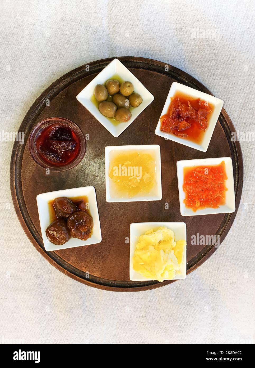 Seven dishes of condiments with jams, olives, and butter arranged on a