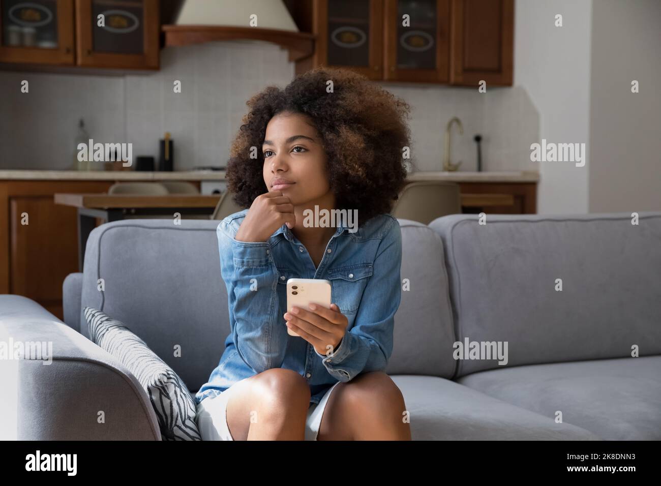 Pretty pensive African teenager girl sit on sofa with smartphone Stock Photo