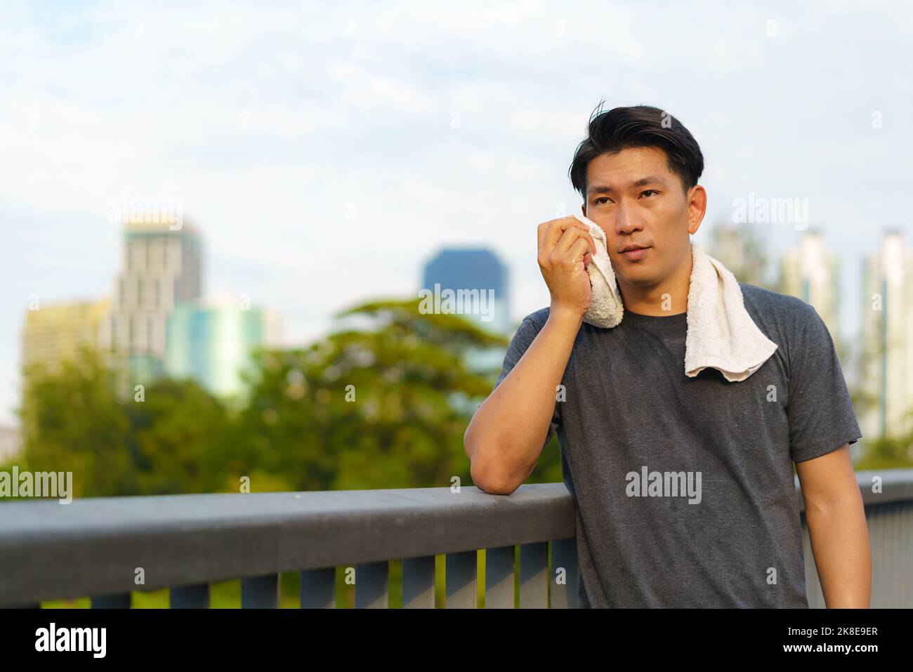 Portrait of Asian young man wearing sportswear, standing and wiping sweat with towel outdoor on walkway with city building background. Stock Photo