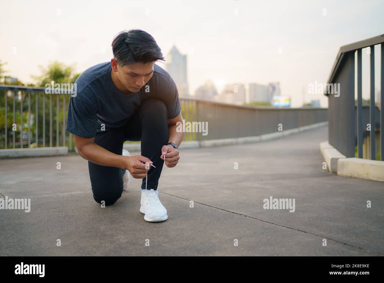 Close-up of Asian man tying shoelaces on white sneakers. Male person squatting on sidewalk. Sunny morning or day. Male going on footpath. Sport and he Stock Photo