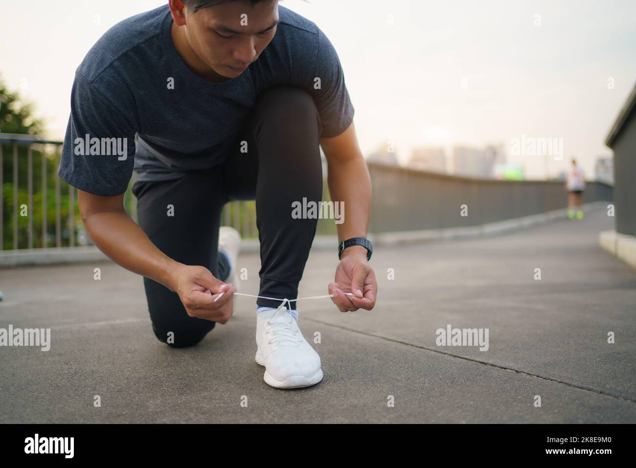 Close-up of Asian man tying shoelaces on white sneakers. Male person squatting on sidewalk. Sunny morning or day. Male going on footpath. Sport and he Stock Photo