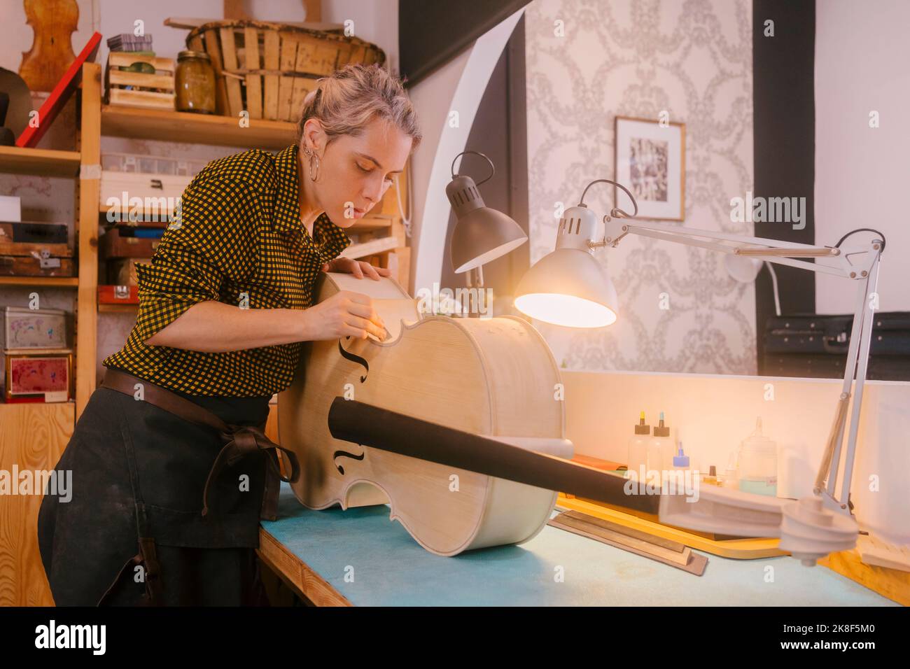 Luthier with hand tool working on double bass standing by desk at