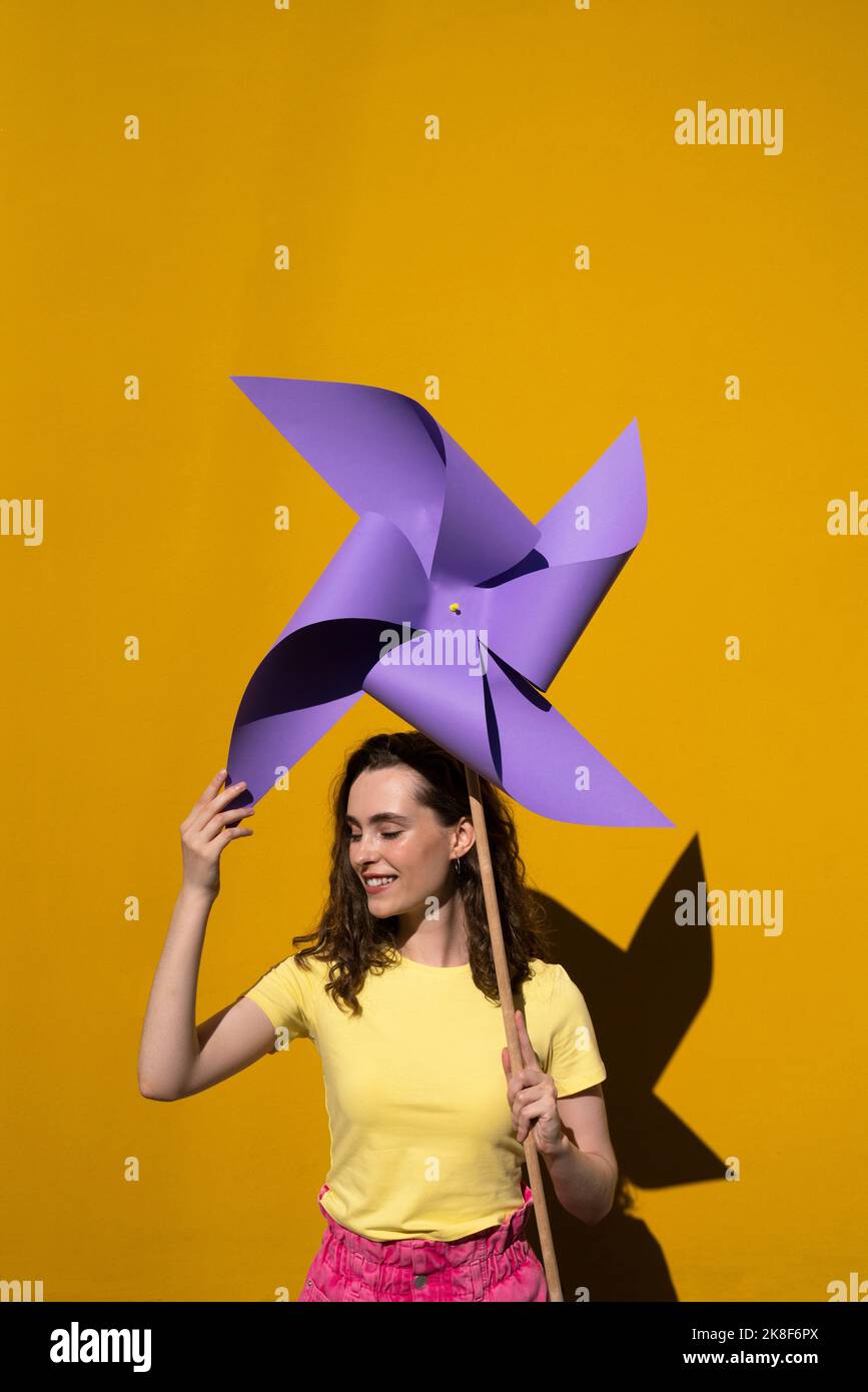 Smiling woman with eyes closed holding pinwheel toy in front of vibrant yellow wall Stock Photo
