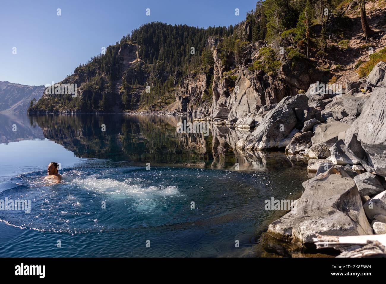 Man going in for a swim in Crater Lake NP Oregon in a beautiful calm afternoon, the Cleetwood Cove trail leads to the Shore and it is one of the only Stock Photo