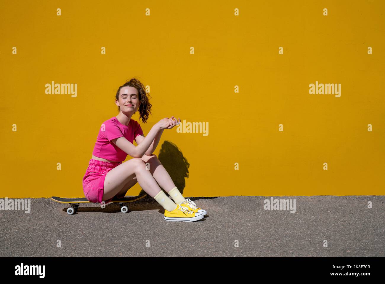 Smiling woman with eyes closed sitting on skateboard in front of yellow wall Stock Photo