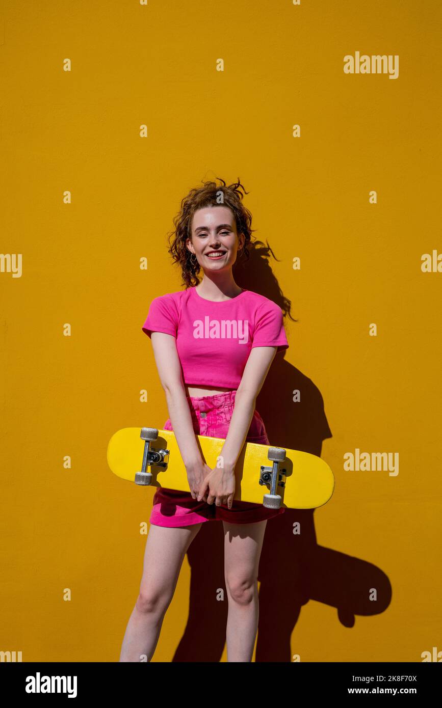 Smiling woman holding skateboard standing in front of vibrant yellow wall Stock Photo