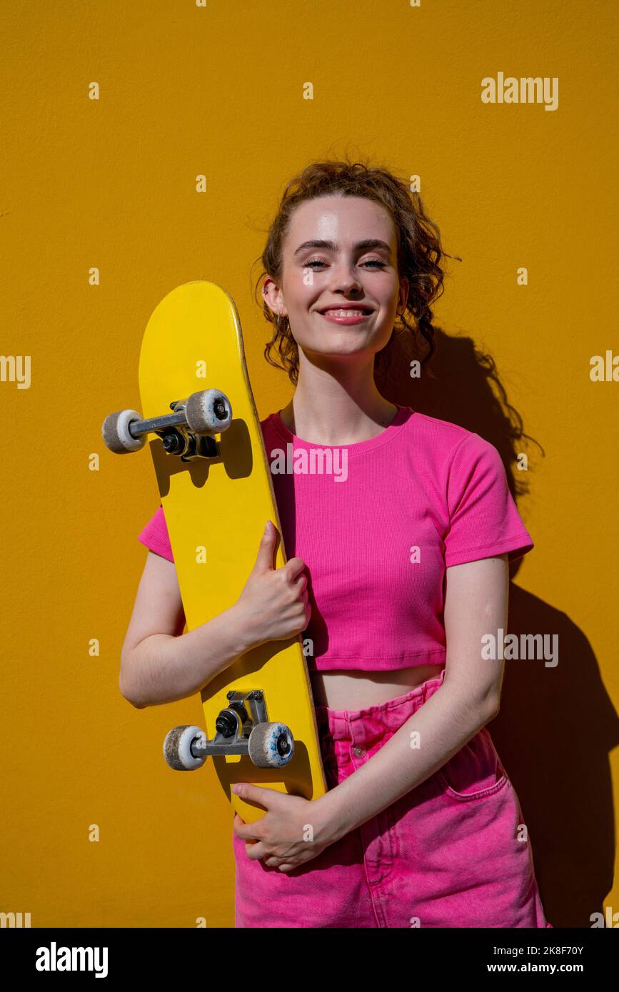 Smiling woman holding yellow skateboard in front of wall Stock Photo