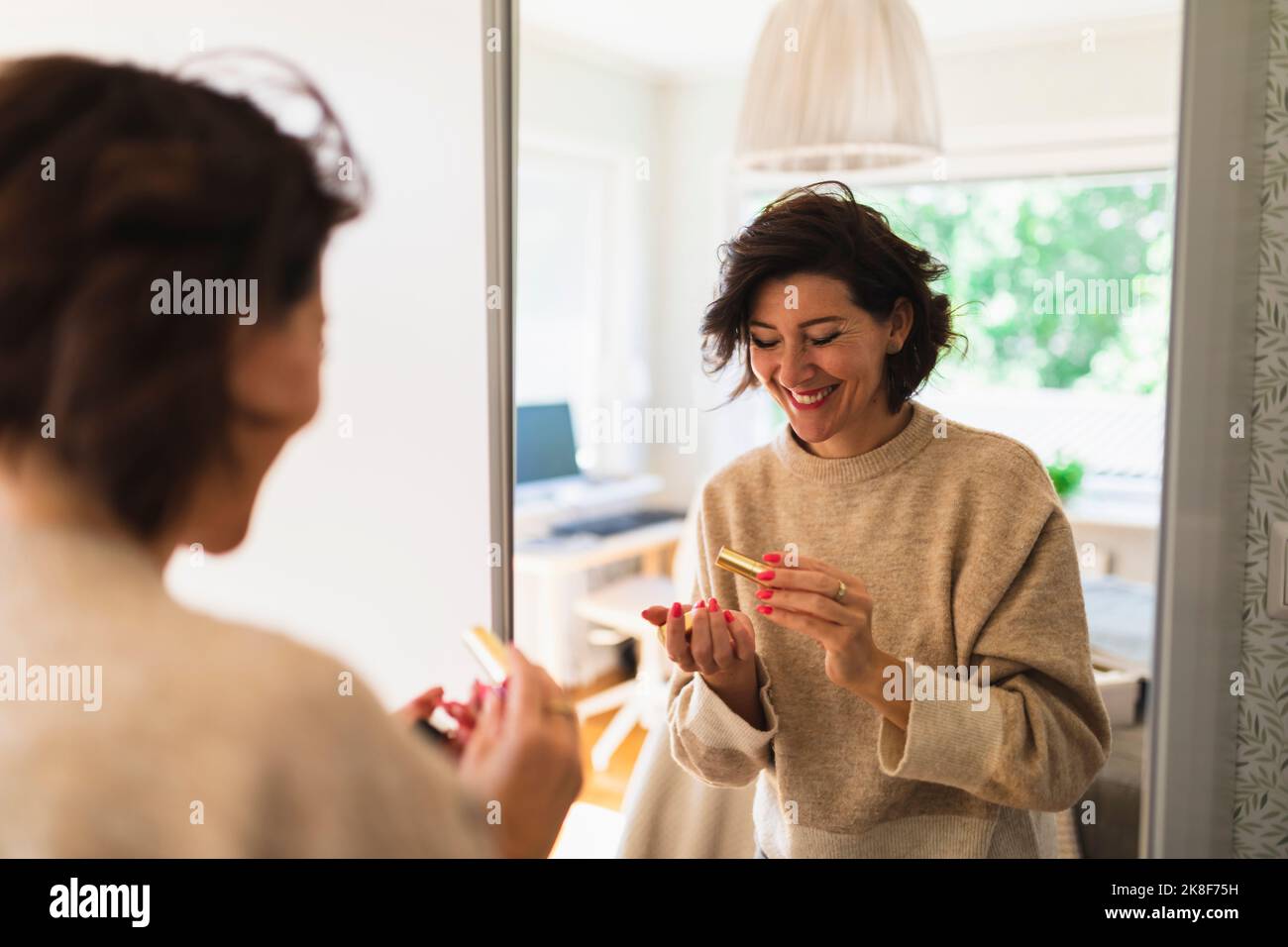 Happy woman holding lipstick in front of mirror at home Stock Photo