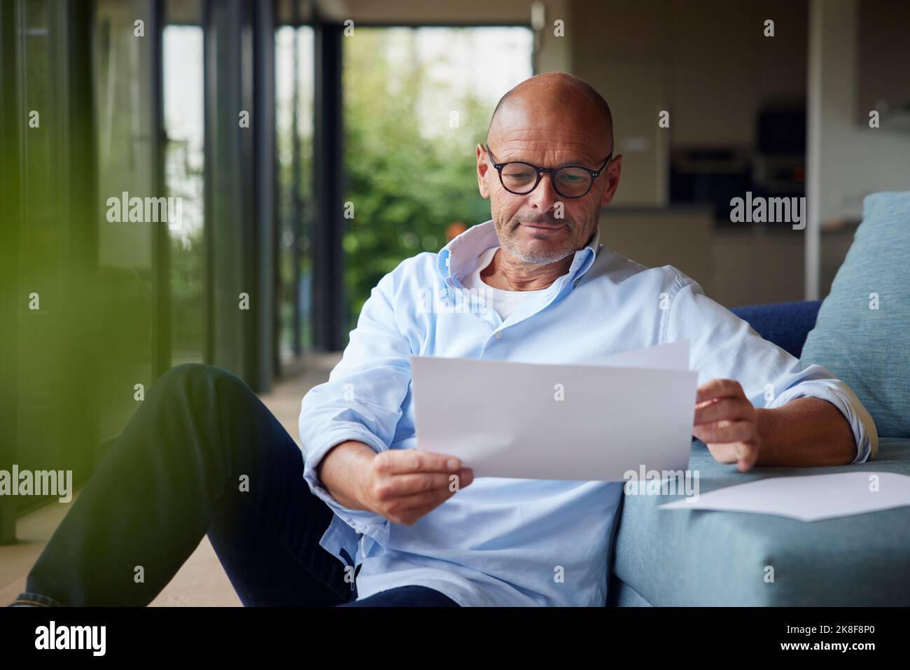 Senior man reading documents sitting by sofa at home Stock Photo