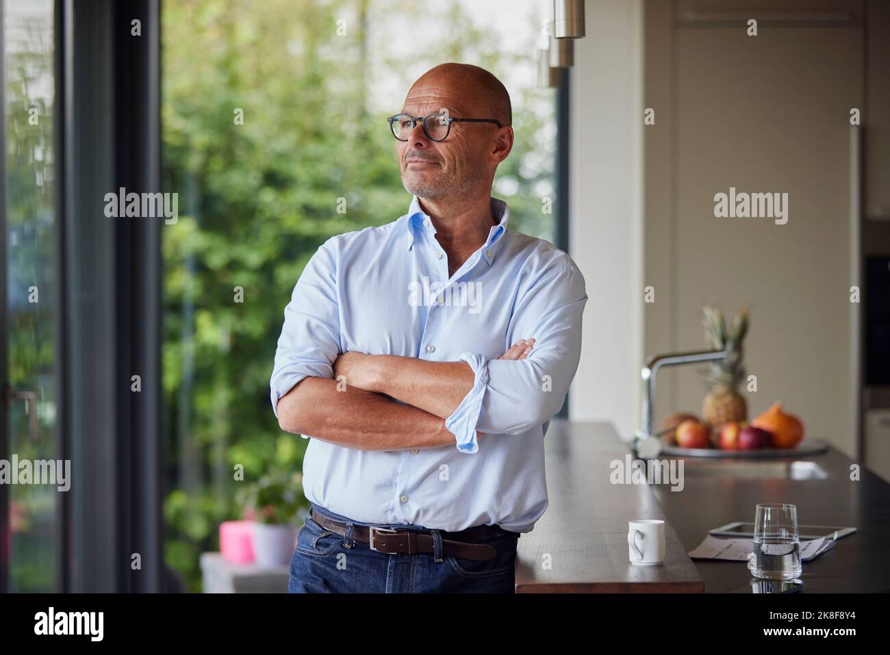 Thoughtful senior man with arms crossed standing by kitchen island at home Stock Photo